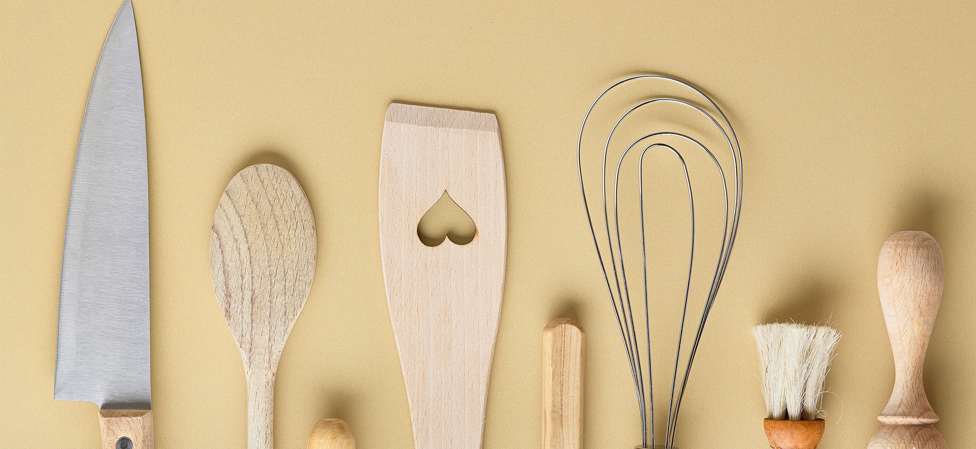 Eine Anordnung von Küchenutensilien auf einem beige Hintergrund. Dazu gehören ein Messer, verschiedene Holzlöffel, ein Rührbesen mit Draht, ein Spatel mit Herzform, sowie eine Bürste und weitere kleine Utensilien.