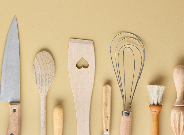 Eine Anordnung von Küchenutensilien auf einem beige Hintergrund. Dazu gehören ein Messer, verschiedene Holzlöffel, ein Rührbesen mit Draht, ein Spatel mit Herzform, sowie eine Bürste und weitere kleine Utensilien.