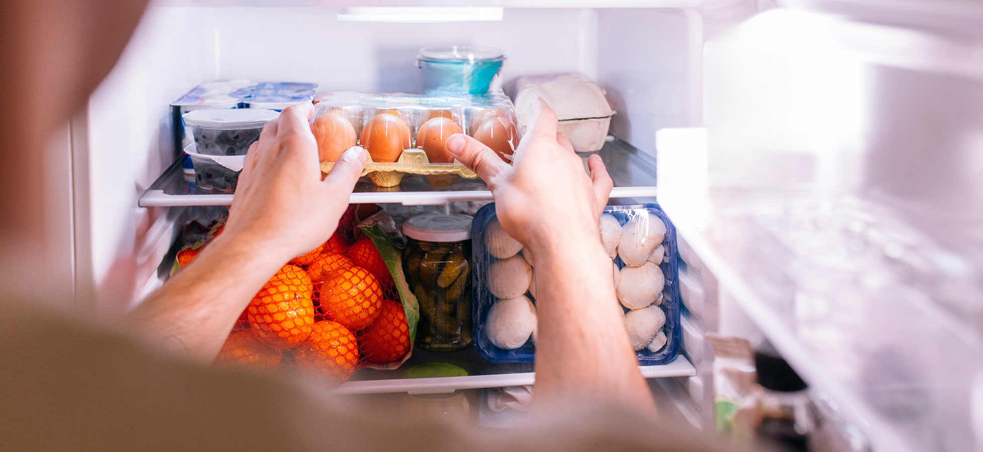 Eine Person greift in einen Kühlschrank und hält eine Packung Eier in den Händen. Im Kühlschrank sind auch orangefarbene Mandarinen und verschiedene Behälter mit Lebensmitteln sichtbar.
