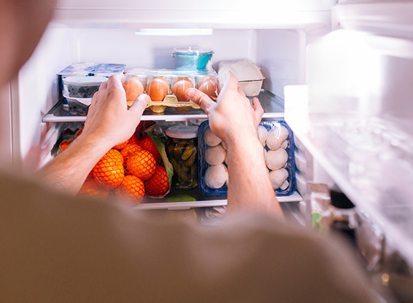 Eine Person steht vor einem offenen Kühlschrank und hält einen Karton mit Eiern in der Hand. Im Kühlschrank sind neben den Eiern auch Orangen und verschiedene Lebensmittelverpackungen sichtbar.