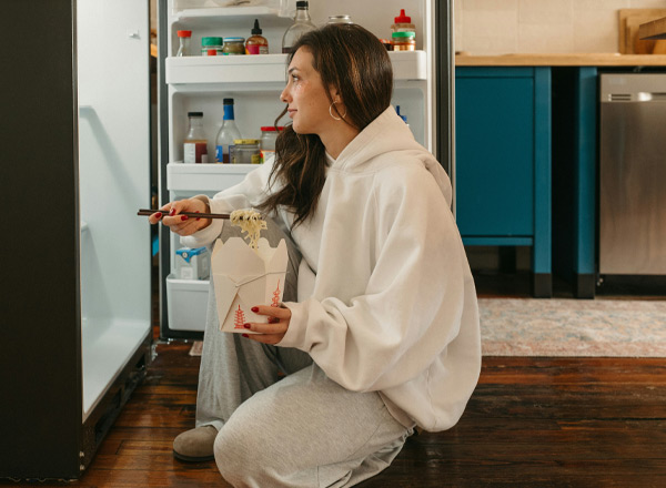 Eine Person sitzt vor einem offenen Kühlschrank, hält eine Gabel und eine Takeaway-Box mit Nudeln. Sie trägt einen grauen Hoodie und schaut nachdenklich in den Kühlschrank, während sie die Nudeln isst. Die Szene vermittelt eine entspannte essensbezogene Stimmung.