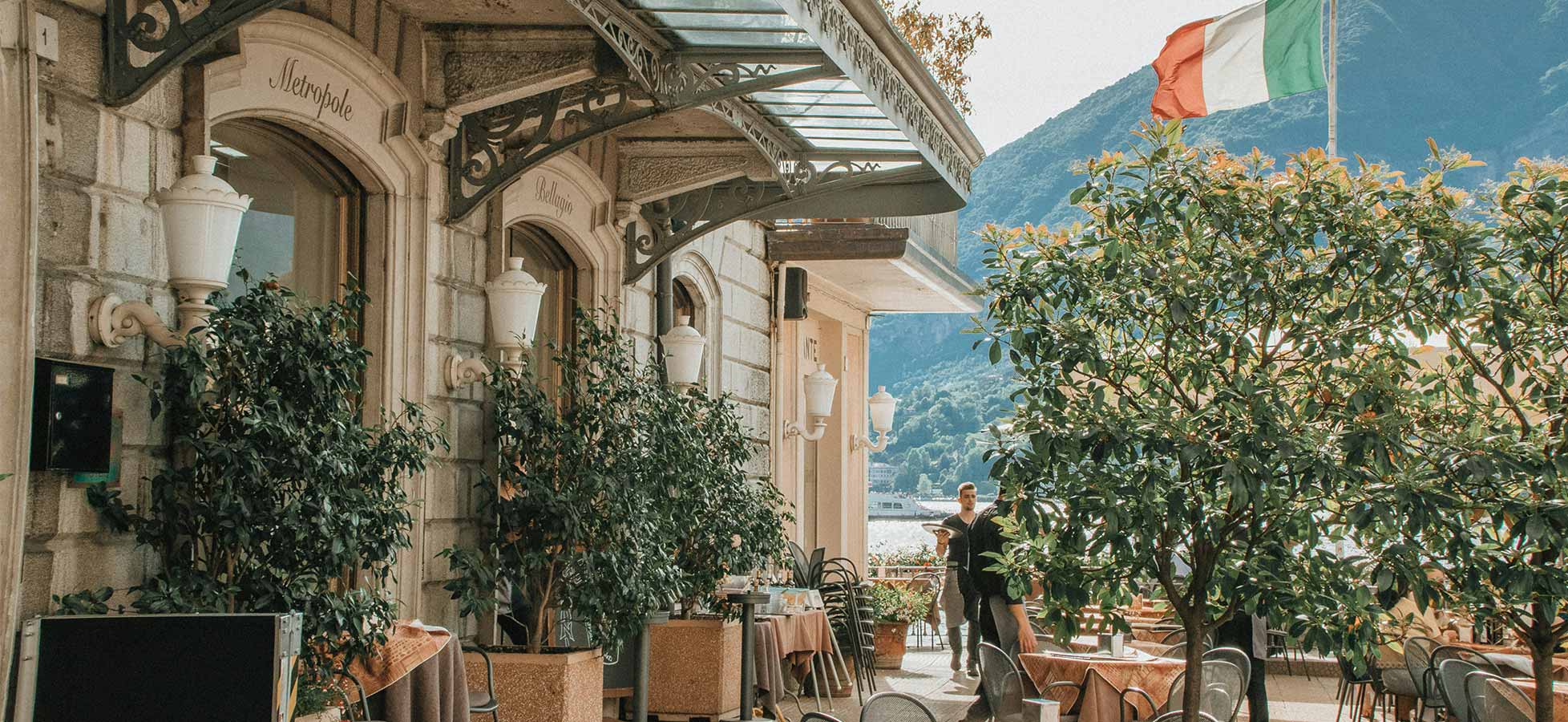 Ein charmantes Restaurant im Freien mit Tischen und Stühlen, umgeben von üppigem Grün. Im Hintergrund sind Berge und eine italienische Flagge sichtbar. Ein Kellner ist dabei, Gäste zu bedienen. Die Atmosphäre ist einladend und entspannend.