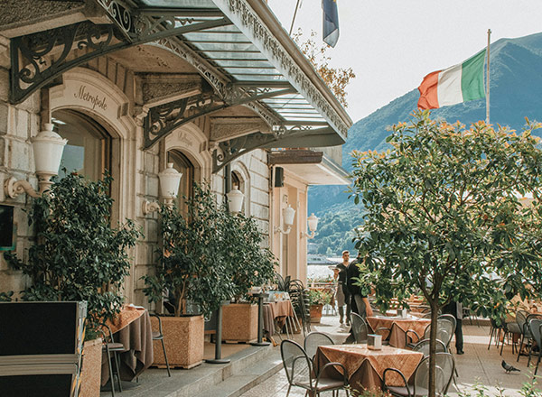 Ein charmantes Restaurant im Freien mit Tischen und Stühlen, umgeben von üppigem Grün. Im Hintergrund sind Berge und eine italienische Flagge sichtbar. Ein Kellner ist dabei, Gäste zu bedienen. Die Atmosphäre ist einladend und entspannend.