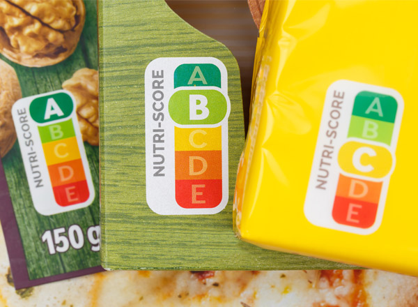 Auf dem Bild sind mehrere Nahrungsmittelverpackungen zu sehen, die den Nutri-Score zeigen. Dieser Score ist eine farbcodierte Kennzeichnung mit den Buchstaben A bis E, die die Nährwertqualität der Produkte bewertet. A steht für die beste und E für die schlechteste Bewertung.