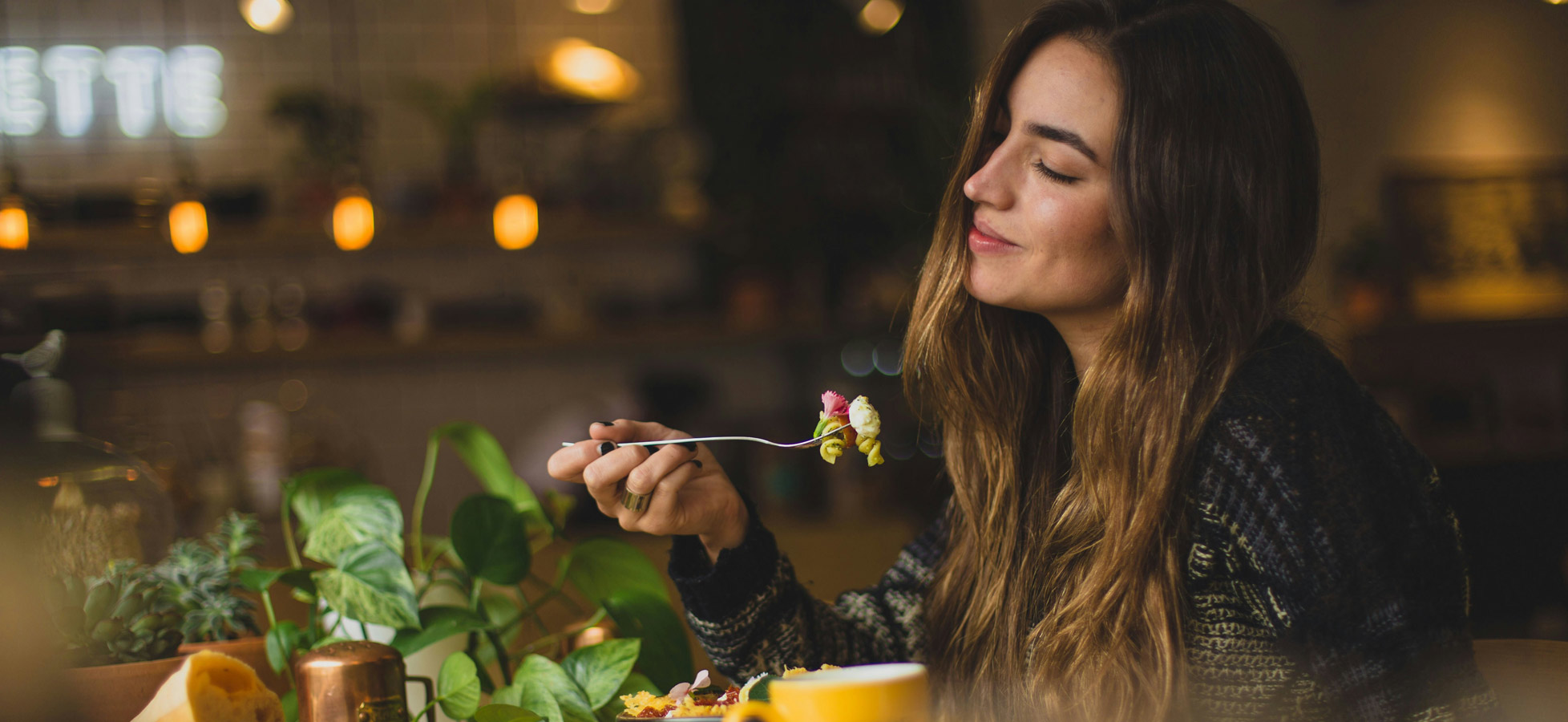 Eine Frau mit langen Haaren lächelt, während sie einen Löffel mit buntem Essen an den Mund führt. Im Hintergrund sind Pflanzen und Lichtquellen sichtbar, die eine gemütliche Atmosphäre erzeugen.
