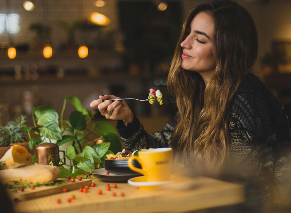 Eine Frau sitzt an einem Tisch und genießt eine Mahlzeit. Sie hält eine Gabel mit buntem Gemüse und lächelt zufrieden. Auf dem Tisch sind gesunde Lebensmittel, eine Tasse und eine Pflanze zu sehen, die eine einladende Atmosphäre schaffen.