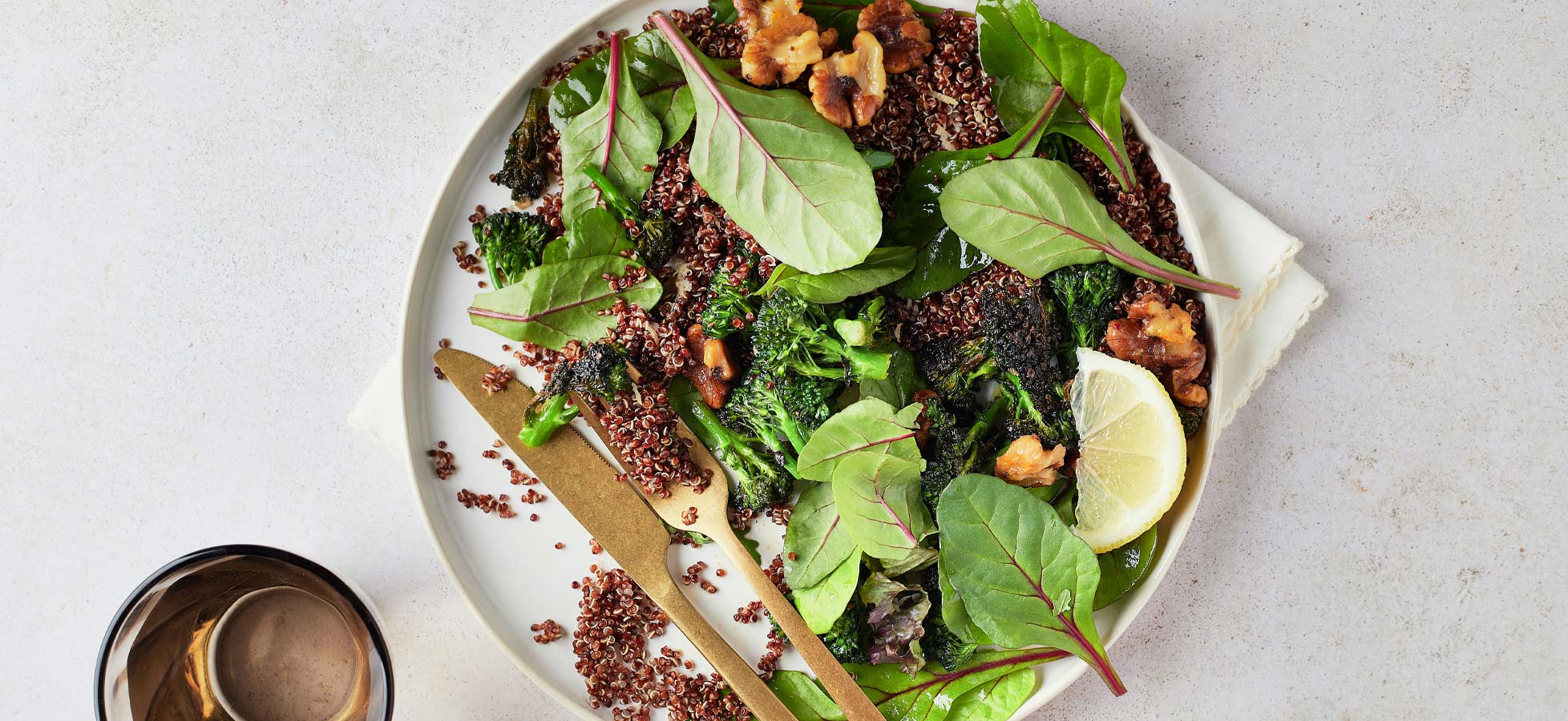 Ein Teller mit einem bunten Salat aus frischem Blattgemüse, Quinoa und Nüssen, garniert mit Zitronenscheiben. Neben dem Teller liegt Besteck, und ein Glas steht daneben. Die Komposition vermittelt einen gesunden, appetitlichen Eindruck.