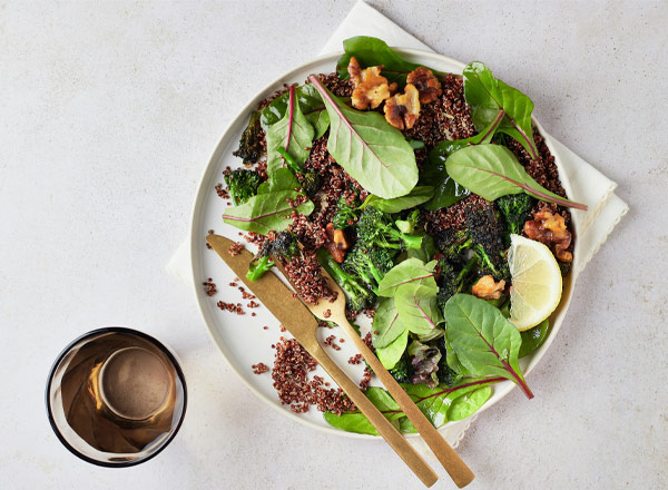 Ein Teller mit einem bunten Salat aus frischem Blattgemüse, Quinoa und Nüssen, garniert mit Zitronenscheiben. Neben dem Teller liegt Besteck, und ein Glas steht daneben. Die Komposition vermittelt einen gesunden, appetitlichen Eindruck.