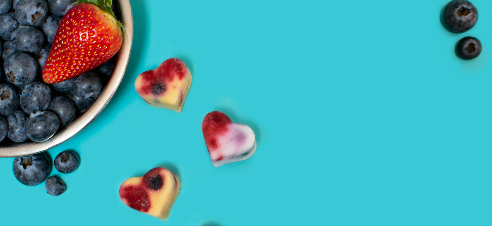 Schüssel mit blauen Heidelbeeren, einer Erdbeere und bunten Herzstückchen aus gefrorenem Fruchtsaft. Der Hintergrund ist leuchtend türkis. Die Anordnung vermittelt eine frische, einladende Atmosphäre von gesunden Snacks.