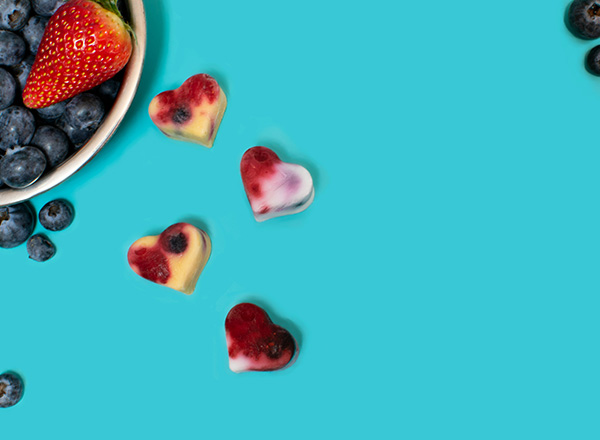 Eine Schale mit Blaubeeren und einer Erdbeere befindet sich auf einem türkisfarbenen Hintergrund. Neben der Schale liegen vier herzförmige, gefrorene Fruchtstückchen in verschiedenen Farben, die aus verschiedenen Beeren bestehen.