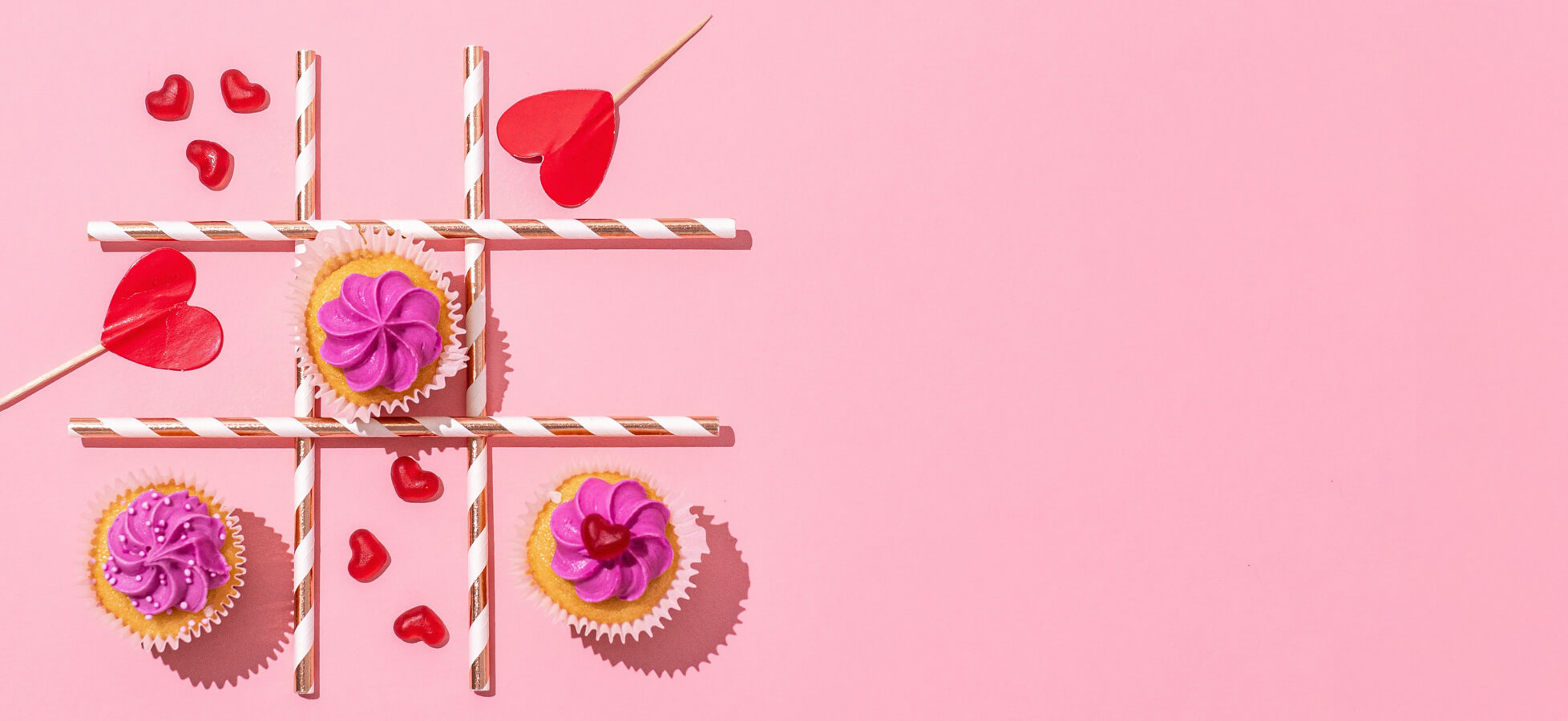 Drei Cupcakes mit lila Cremetopping sind auf einem rosa Hintergrund angeordnet. Zwischen ihnen liegen gestreifte Strohhalme in Form eines Kreutzspiels. Rote Herzchen aus Zucker sind verstreut, was eine verspielte und festliche Stimmung erzeugt.