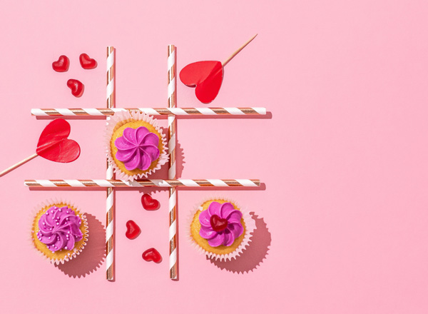Drei Cupcakes mit buntem Zuckerguss befinden sich auf einem rosa Hintergrund, angeordnet wie ein Spielbrett. Rote Herzen und bunte Strohhalme ergänzen das Bild, das eine verspielte und festliche Atmosphäre vermittelt.