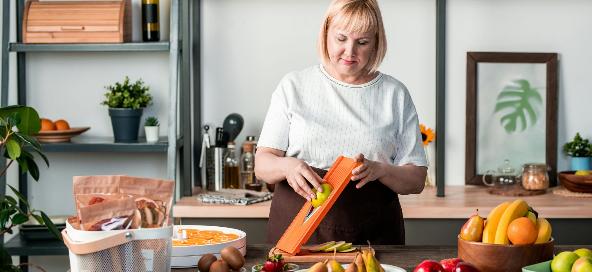 Eine Frau mit blonden Haaren schneidet mit einem orangefarbenen Küchenwerkzeug einen grünen Apfel. Vor ihr liegen verschiedene Früchte, darunter Kiwis, Äpfel und Bananen, auf einem Holztisch. Im Hintergrund sind Regale mit Obst und Küchenutensilien sichtbar.