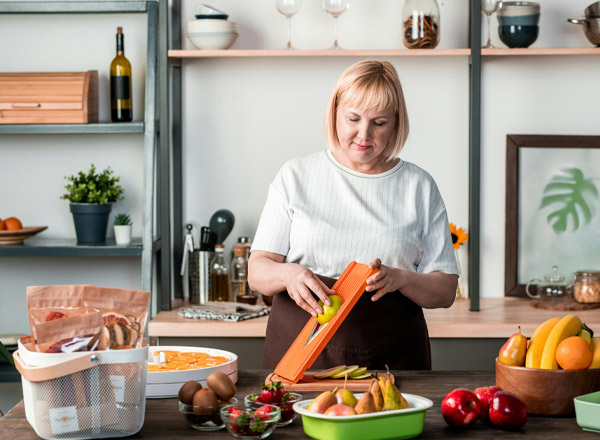 Eine Frau mit blonden Haaren steht an einem Tisch und schneidet Gemüse mit einem Küchenwerkzeug. Um sie herum liegen frisches Obst und Gemüse in verschiedenen Schalen. Im Hintergrund sind Regale mit Kochutensilien und dekorativen Elementen sichtbar.