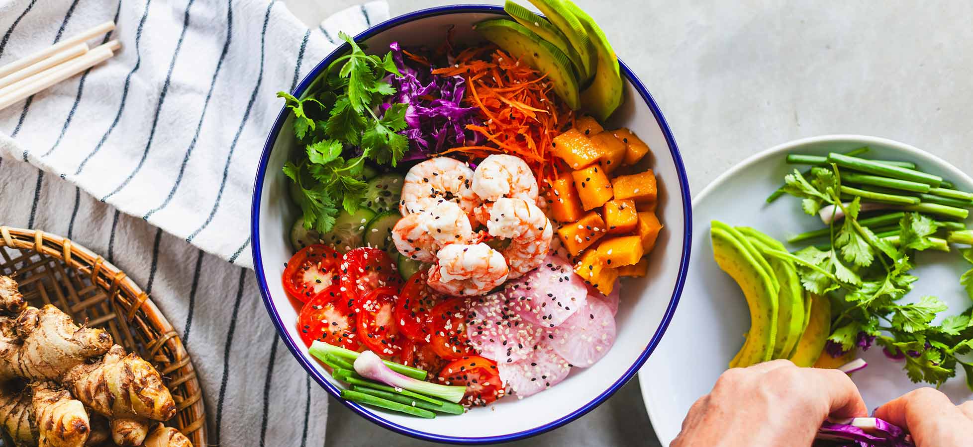 Eine Schüssel mit buntem Salat, darunter Garnelen, Karotten, Avocado, Tomaten und frische Kräuter. Neben der Schüssel befinden sich frische Zutaten wie Ingwer und Schnittlauch sowie ein Teller mit zusätzlichen Kräutern. Das Bild zeigt eine appetitliche, gesunde Mahlzeit.