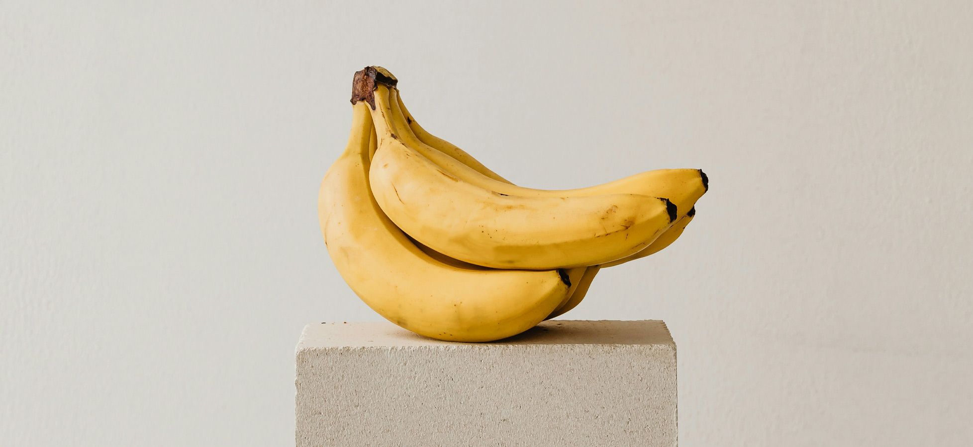 Ein Paar reife, gelbe Bananen liegt auf einem schlichten, hellen Podest. Der Hintergrund ist neutral und trägt zur minimalistischen Darstellung der Bananen bei.