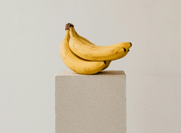 Ein Paar reife, gelbe Bananen liegt auf einem schlichten, hellen Podest. Der Hintergrund ist neutral und trägt zur minimalistischen Darstellung der Bananen bei.