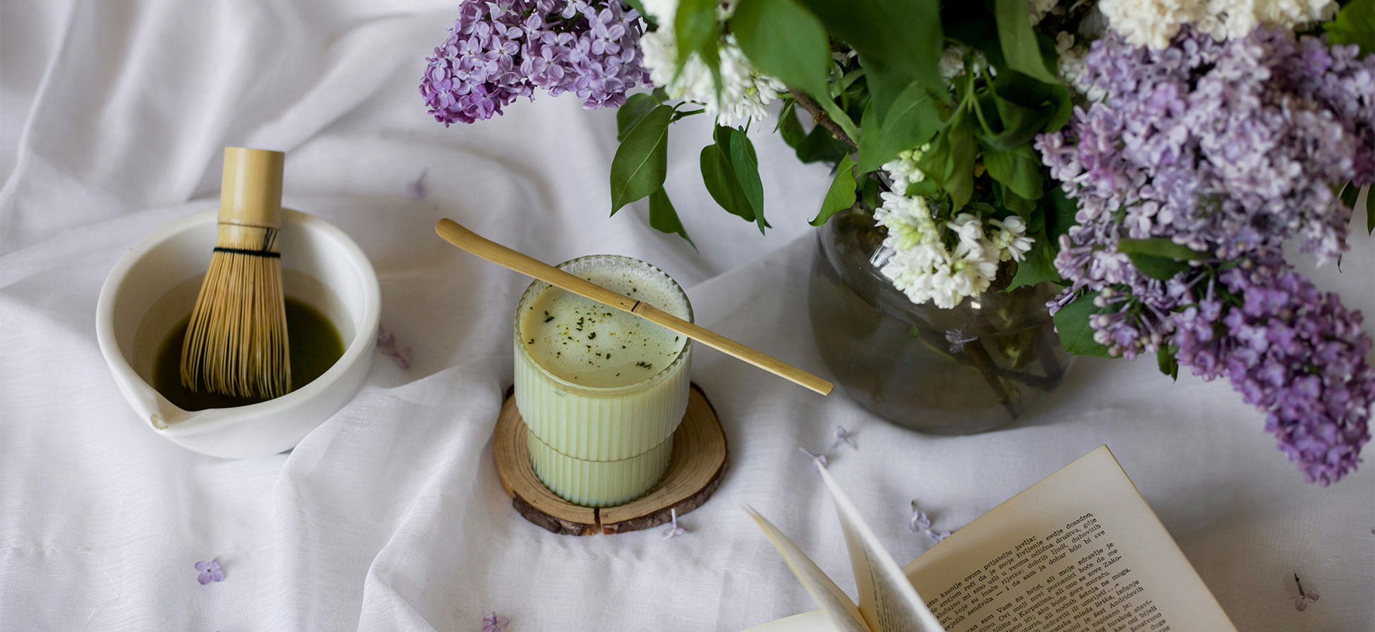 Ein Tee-Set mit einem Schalenmixbesen und einer Tasse mit grünem Tee auf einem Holzuntersetzer. Neben der Tasse steht eine Vase mit frischen, bunten Fliederblüten. Ein aufgeschlagenes Buch liegt im Vordergrund auf einem weißen Tuch.