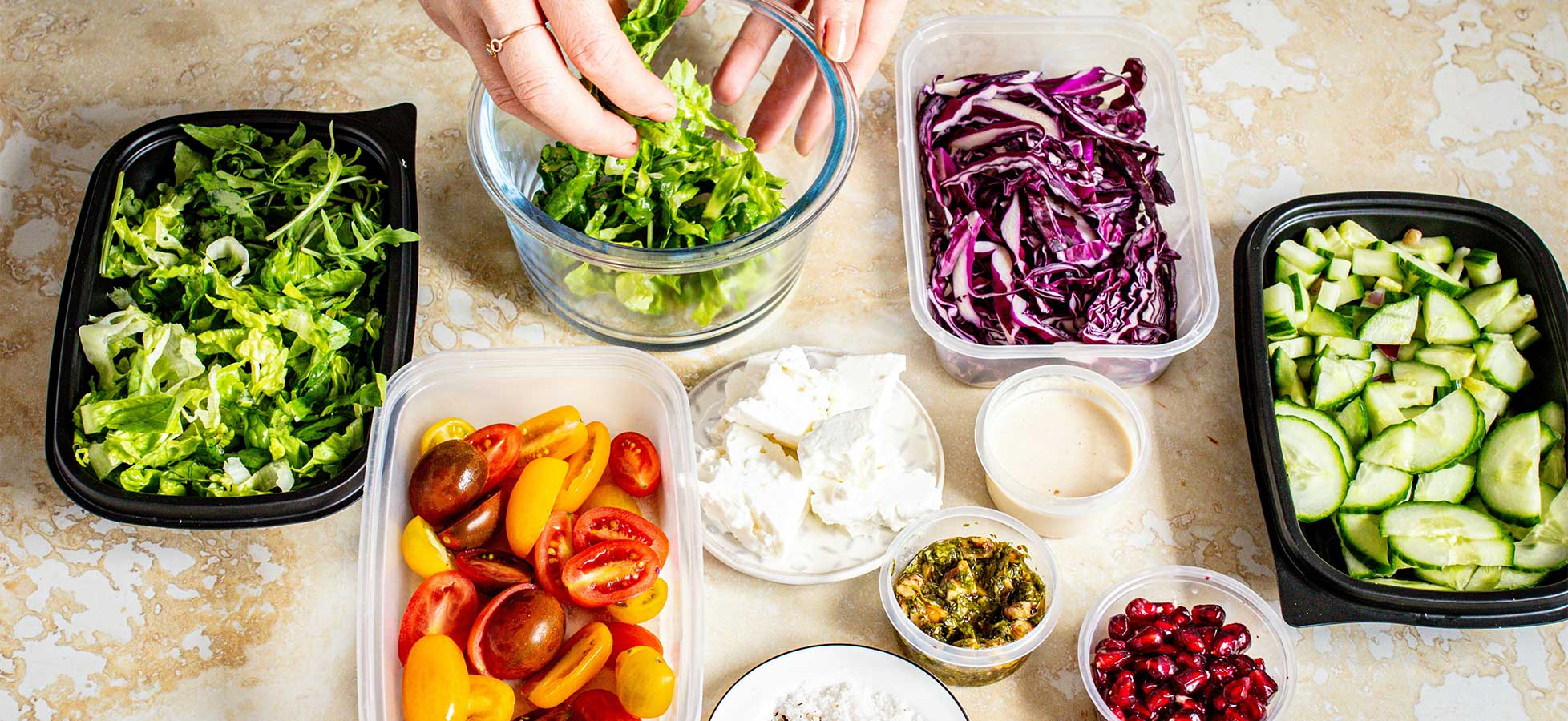 Eine Person bereitet einen Salat vor. Verschiedene frische Zutaten sind auf einem Tisch angeordnet: grüne Blattsalate, rote Kohlsalat, Gurken, Kirschtomaten, Dip und Granatapfelkerne. Die Person fügt Salat zu einer Schüssel hinzu.
