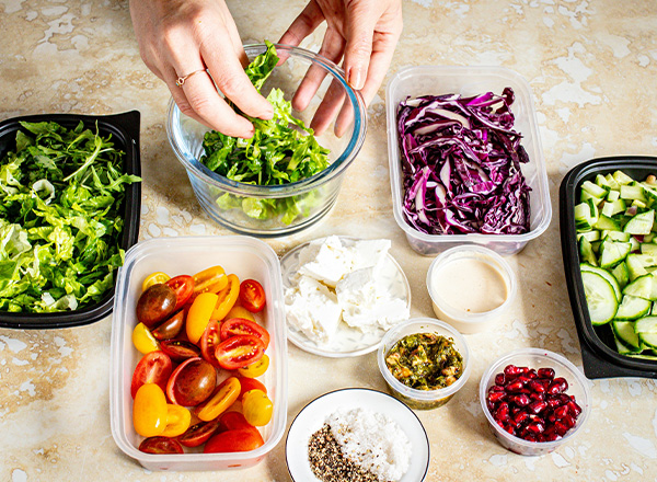 Eine Person greift in eine Schüssel mit frischem, grünen Salat. Rundherum sind verschiedene Zutaten wie rote und gelbe Tomaten, Feta-Käse, Kräuterpaste und Sesam in Schalen angeordnet. Der Hintergrund ist eine helle, marmorierte Oberfläche.