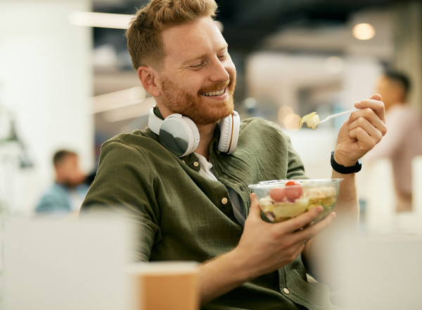 Ein lächelnder Mann mit roten Haaren und Bart sitzt in einem modernen Raum, trägt Kopfhörer und hält eine Schüssel mit Salat und frischem Obst. Er scheint den Moment zu genießen, während er mit einer Gabel in der Schüssel isst.