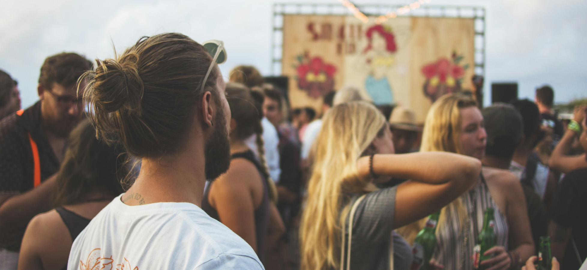 Eine Gruppe von Menschen steht auf einem Festivalgelände. Die Atmosphäre ist lebhaft, und die Teilnehmer, einige mit Getränken in der Hand, blicken auf eine große Bühne im Hintergrund, die mit einem bunten Banner dekoriert ist.