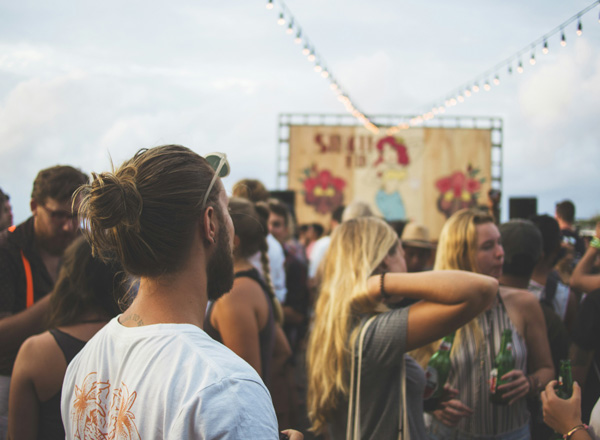 Eine Gruppe von Menschen steht auf einem Festival, einige halten Getränke in der Hand. Im Hintergrund ist eine große Leinwand mit einer bunten Illustration zu sehen. Lichterketten hängen über den Köpfen der Menschen, und die Atmosphäre wirkt lebhaft und fröhlich.