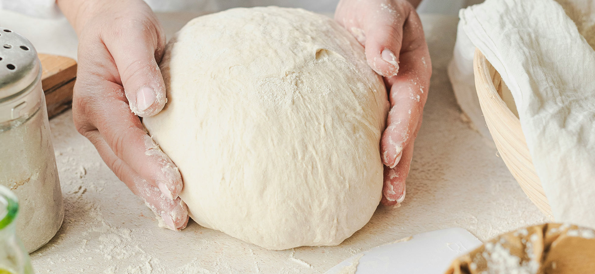 Eine Person hält einen runden Stück Teig in beiden Händen, umgeben von etwas Mehl auf einer Arbeitsfläche. Im Hintergrund sind weitere Küchenelemente wie ein Korb und ein Salzgefäß zu sehen.