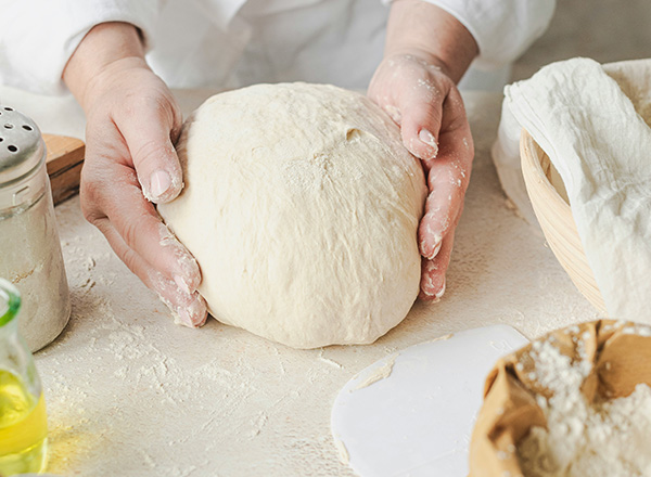 Eine Person hält einen großen, runden Teigball in den Händen. Der Tisch ist leicht mit Mehl bestäubt, und im Hintergrund sind weitere Küchenutensilien sichtbar, die zum Backen verwendet werden.