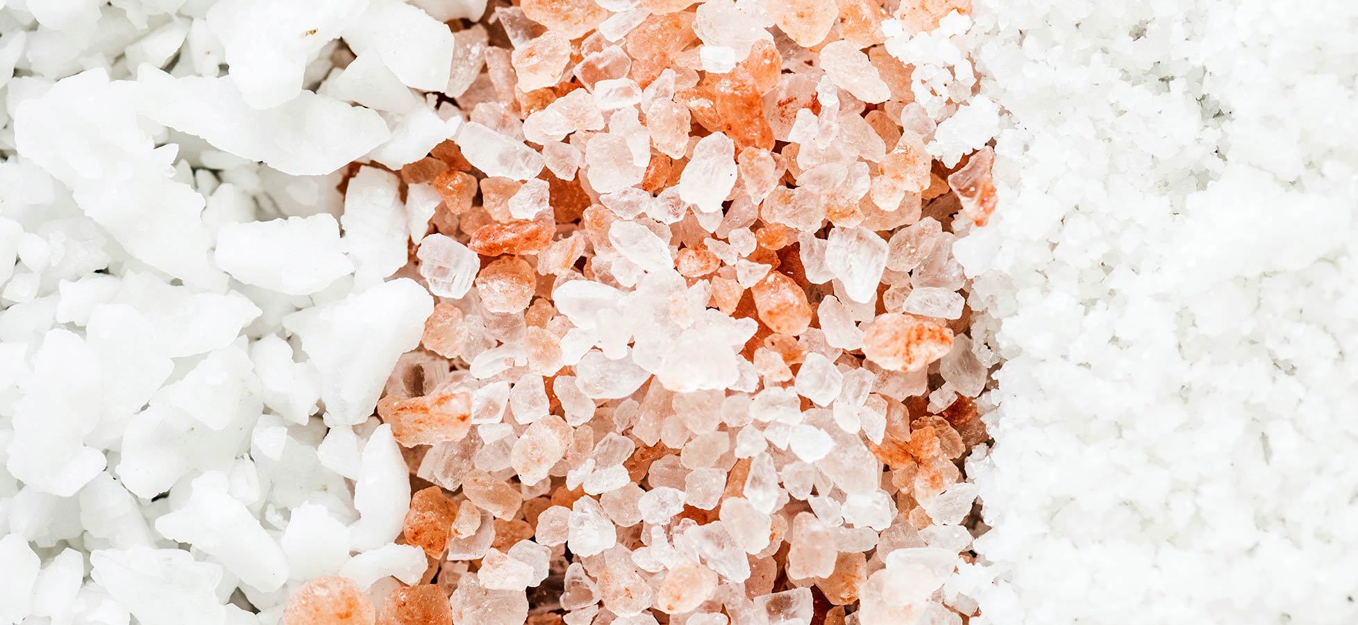 Das Bild zeigt verschiedene Salze: auf der linken Seite grobes, weißes Salz, in der Mitte rosa Salz und rechts feines, weißes Salz. Die Salze sind unterschiedlich in Farbe und Struktur, was die Vielfalt von Salzarten veranschaulicht.