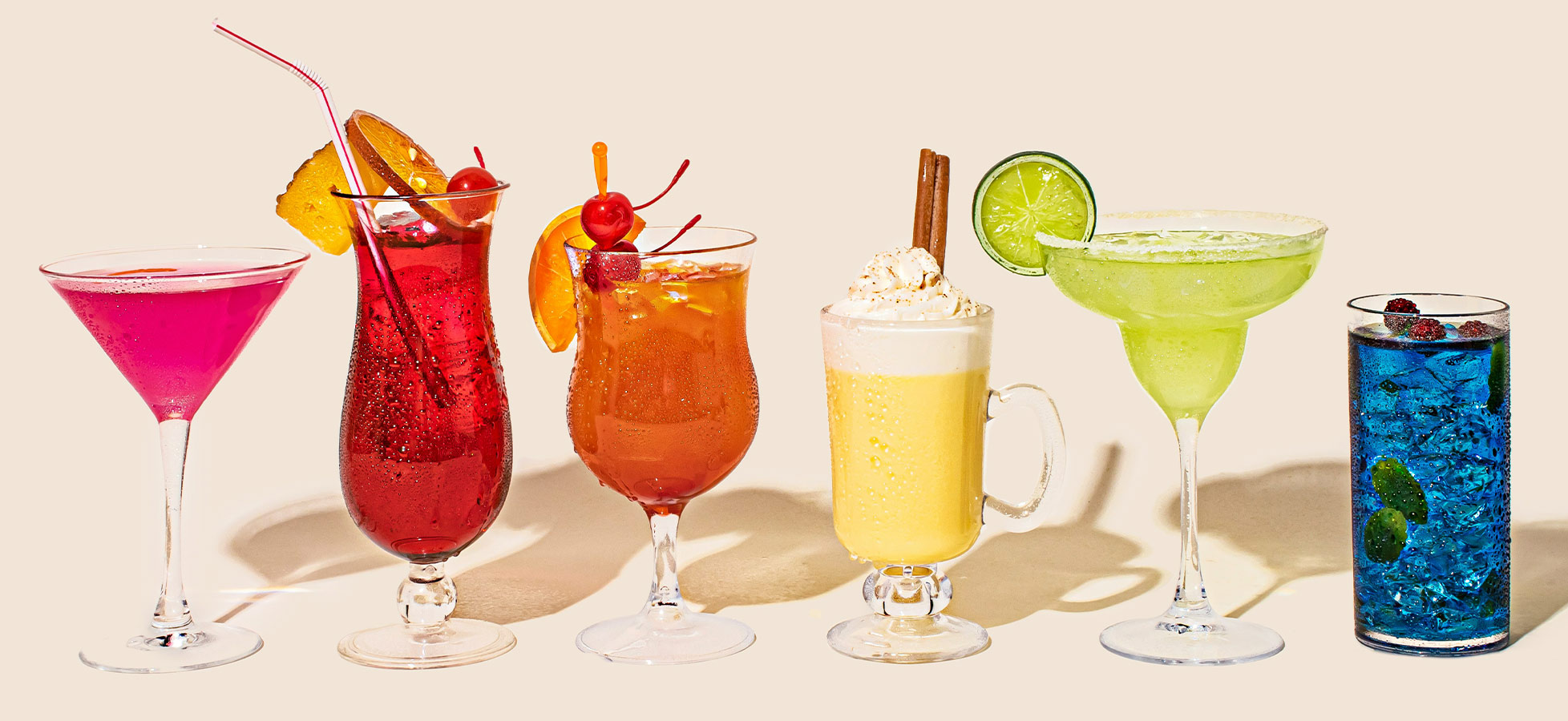 Das Bild zeigt eine Reihe von sechs verschiedenen Cocktails in dekorativen Gläsern. Die Getränke sind bunt, inklusive roter, orangefarbener, gelber, grüner und blauer Farben, und einige enthalten Garnierungen wie Früchte und Sahne. Sie sind auf einem neutralen Hintergrund arrangiert.