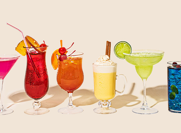 Das Bild zeigt eine Reihe von sechs verschiedenen Cocktails in dekorativen Gläsern. Die Getränke sind bunt, inklusive roter, orangefarbener, gelber, grüner und blauer Farben, und einige enthalten Garnierungen wie Früchte und Sahne. Sie sind auf einem neutralen Hintergrund arrangiert.