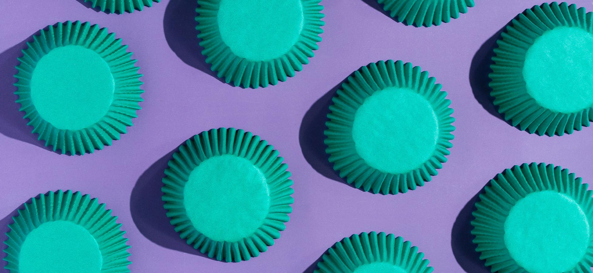 Eine Fläche mit wiederholt angeordneten, grünen Backförmchen auf lila Hintergrund. Die Förmchen sind rund mit gewelltem Rand und gleichmäßig verteilt, wodurch ein ansprechendes Muster entsteht.