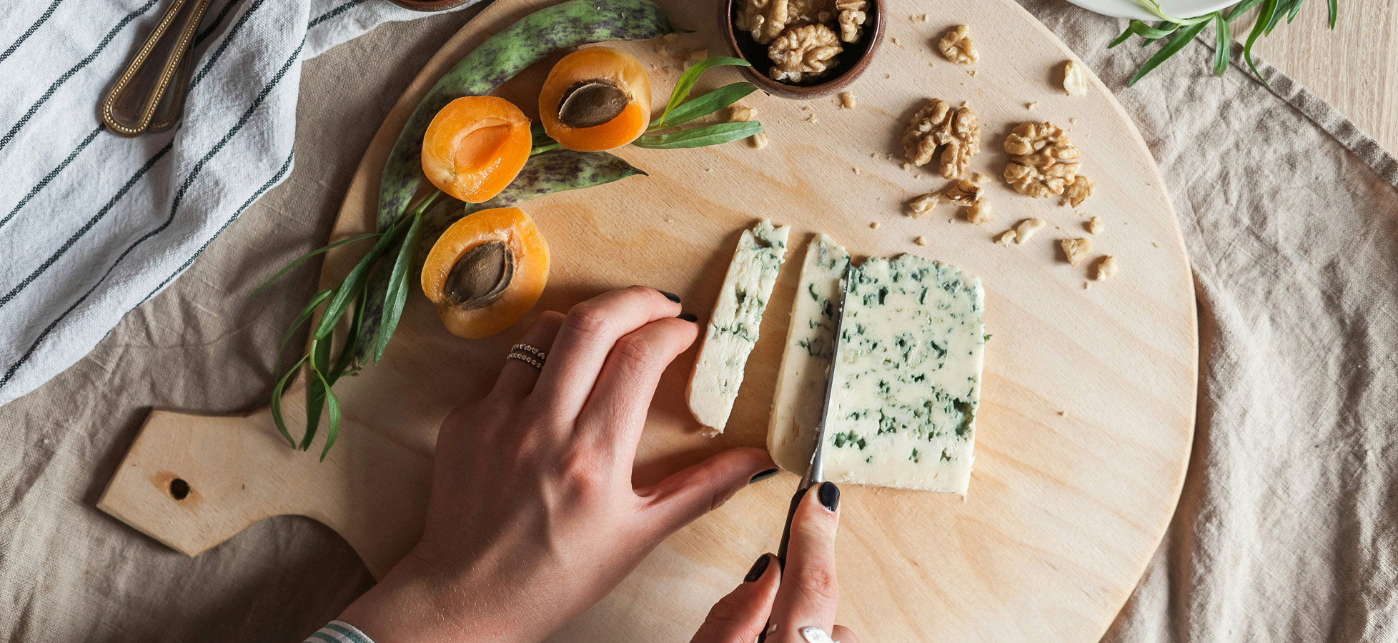 Eine Hand schneidet einen Stück Käse auf einem Holzbrett. Um den Käse liegen frische Aprikosenhälften, Walnüsse und Kräuter. Im Hintergrund ist ein gestreifter Geschirrtuch sichtbar.