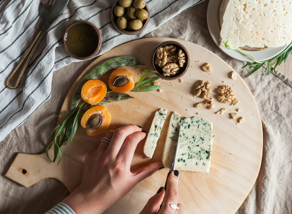 Eine Hand schneidet Käse auf einem runden Holzbrett. Neben dem Käse liegen Aprikosen, Oliven und Walnüsse. Im Hintergrund sind einige Zutaten und eine Serviette sichtbar. Die Szene vermittelt eine einladende Atmosphäre für ein gemeinsames Essen.