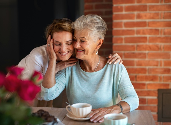 Zwei Frauen sitzen zusammen an einem Tisch, lächeln und genießen einen Moment miteinander. Die Jüngere umarmt die Ältere liebevoll, während eine Tasse Kaffee zwischen ihnen steht. Im Hintergrund sind rosa Blumen sichtbar. Es herrscht eine freundliche und herzliche Atmosphäre.