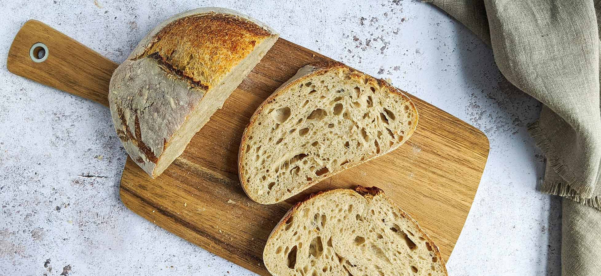 Auf dem Holzbrett liegt ein frisch gebackenes Brot. Ein Teil des Brotes ist aufgeschnitten, und die strukturierte Krume ist sichtbar. Der Hintergrund ist neutral und hebt das Brot hervor.