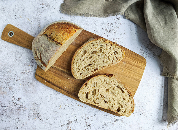 Auf einem Holzbrett liegt ein Laib Brot, das teilweise aufgeschnitten ist. Zwei dicke Scheiben des Brotes sind neben dem Laib angeordnet, und die krustige Oberfläche ist gut sichtbar. Im Hintergrund ist ein Tuch zu sehen.