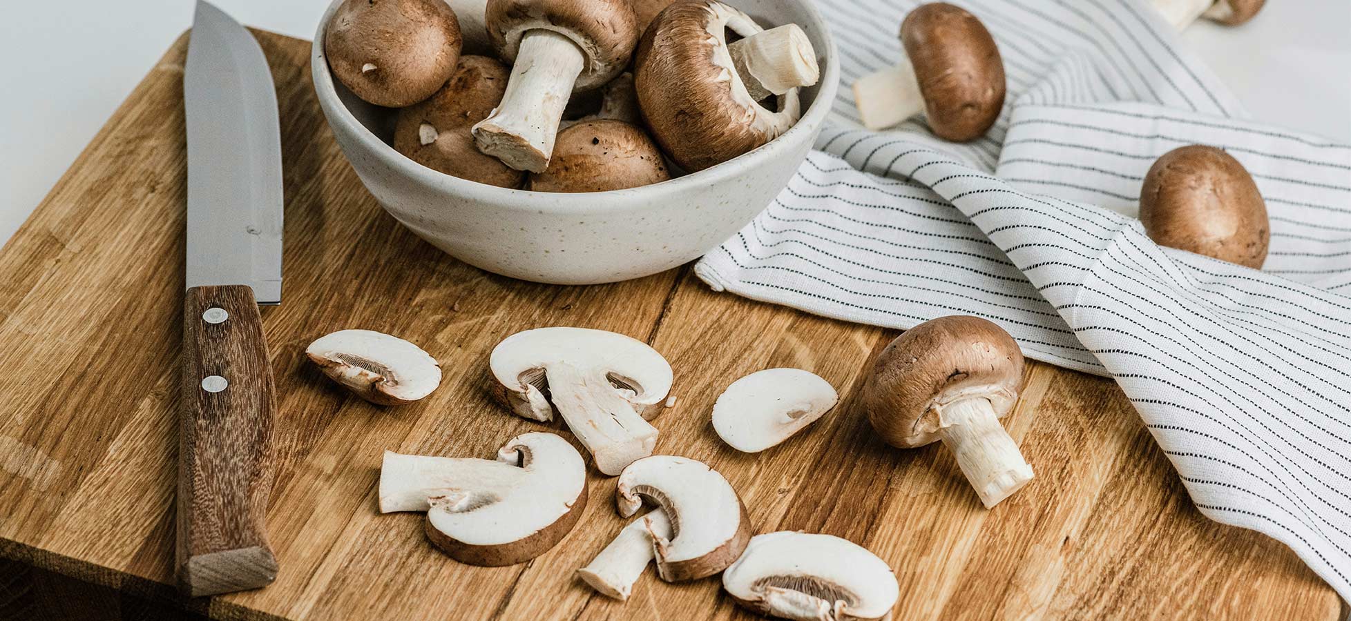 Ein Holztisch mit einem Schälmesser und einem weißen Teller voller brauner Champignons. Einige Champignons sind in Scheiben geschnitten und liegen auf dem Tisch. Ein gestreifter Küchenhandtuch liegt neben dem Teller.