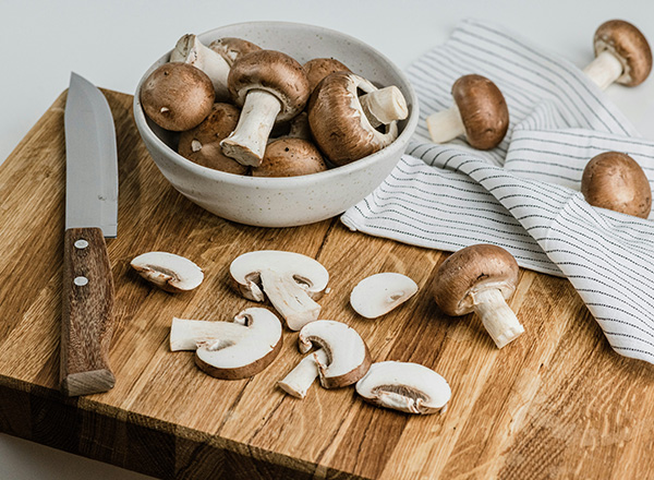 Auf einem Holzbrett liegen geschnittene Champignons neben einer Schüssel mit ganzen Champignons. Ein scharfes Küchenmesser liegt ebenfalls auf dem Brett. Im Hintergrund sind gestreifte Küchentücher zu sehen.