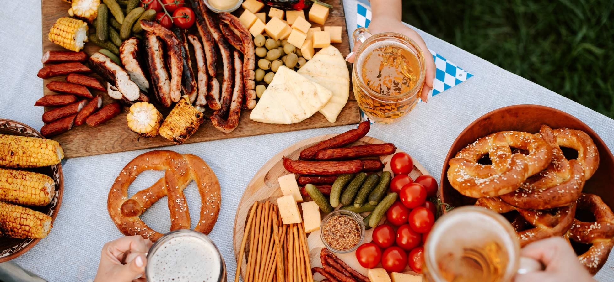 Ein reich gedeckter Tisch mit verschiedenen Grillgerichten wie Würstchen, gegrilltem Mais und Fleisch, sowie einer Auswahl an Käse, Oliven und Gemüse. Dazu werden Brezeln und Bier serviert. Die Atmosphäre wirkt festlich und einladend.
