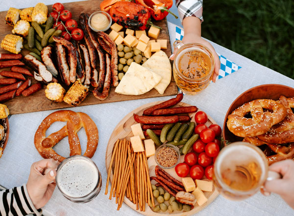 Ein reich gedeckter Tisch mit verschiedenen Grillgerichten wie Würstchen, gegrilltem Mais und Fleisch, sowie einer Auswahl an Käse, Oliven und Gemüse. Dazu werden Brezeln und Bier serviert. Die Atmosphäre wirkt festlich und einladend.
