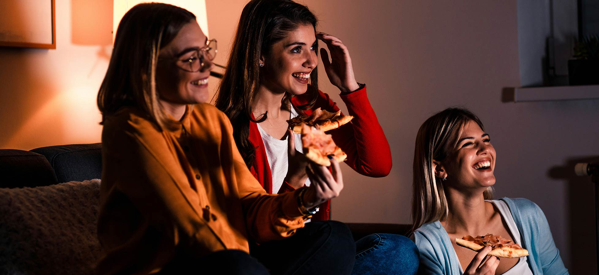Drei Frauen sitzen auf einem Sofa und lachen, während sie Pizza essen. Die Atmosphäre ist gemütlich und entspannt, mit warmem Licht im Hintergrund, das eine freundliche Stimmung schafft. Ihre Freude und das Teilen des Moments sind deutlich spürbar.