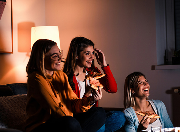 Drei Frauen sitzen in einem gemütlichen Raum und genießen Pizza. Sie lachen und unterhalten sich, während eine sanfte Lichtquelle im Hintergrund eine angenehme Atmosphäre schafft.