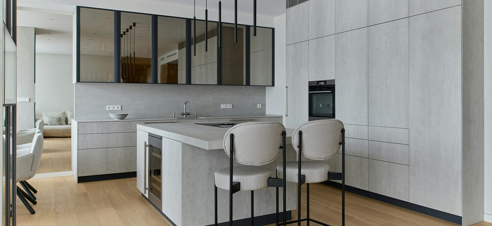 Moderne Küche mit einer großen, grauen Kücheninsel in der Mitte. Es gibt zwei hohe, gepolsterte Stühle und eine stilvolle Gestaltung mit Glasfronten. Die Wände sind in einem hellen Grau gehalten, und der Boden ist aus Holz. Deckenlampen hängen über der Insel.
