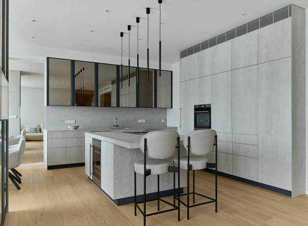 Moderne Küche mit einer großen, grauen Kücheninsel in der Mitte. Es gibt zwei hohe, gepolsterte Stühle und eine stilvolle Gestaltung mit Glasfronten. Die Wände sind in einem hellen Grau gehalten, und der Boden ist aus Holz. Deckenlampen hängen über der Insel.