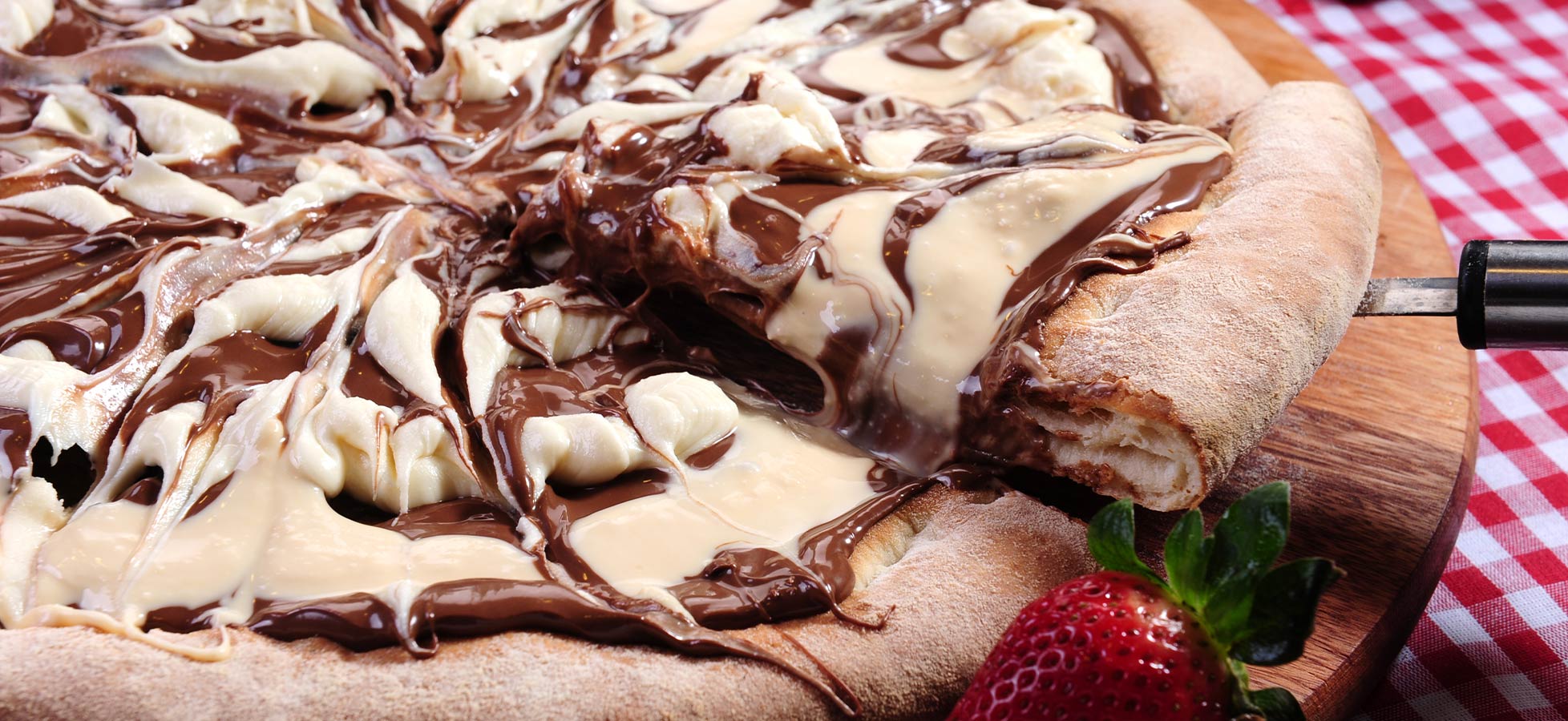 Die Abbildung zeigt eine Pizza mit einer reichhaltigen Schokoladensoße, die kunstvoll mit weißer Schokolade marmoriert ist. Ein Stück der Pizza ist herausgenommen und ein Erdbeerstück liegt daneben. Der Untergrund ist ein Holzbrett.