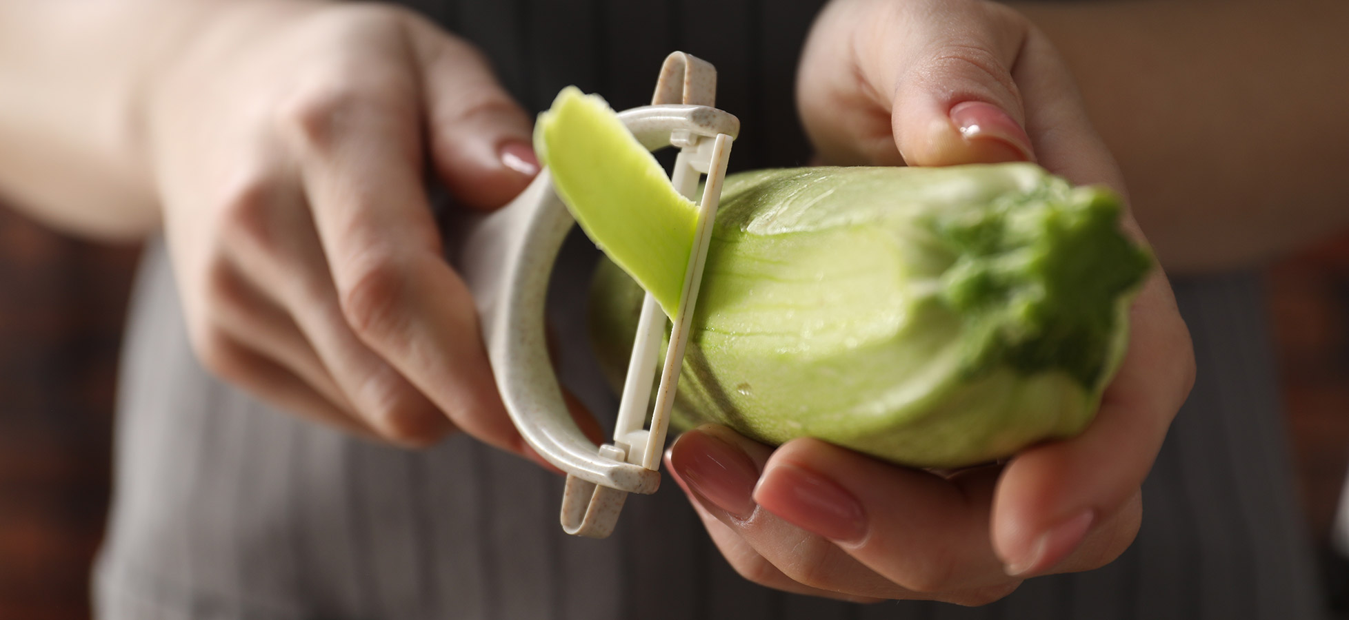 Eine Hand schält mit einem Gemüse Schäler einen grünen Salat. Der Schäler entfernt die äußere Schicht des Salats, während eine weitere Hand das Gemüse hält. Die Szene ist nah aufgenommen und zeigt den Vorgang des Schälens deutlich.