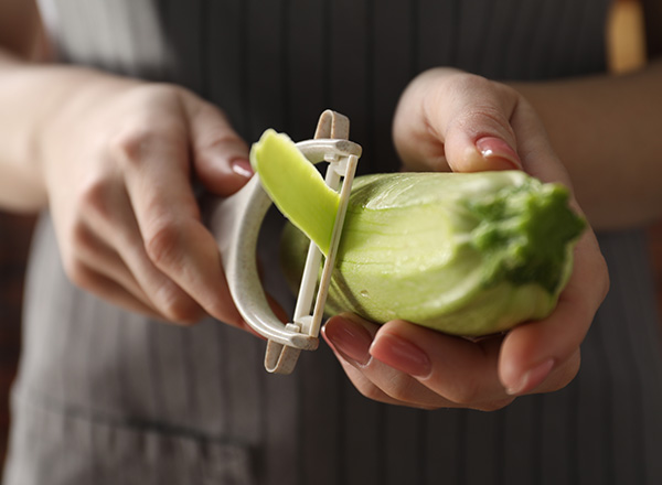 Eine Person schält mit einem Gemüseschäler einen Zucchini. Die Hände sind gut sichtbar, während die Spitze des Schälers in Kontakt mit dem Gemüse ist. Der Zucchini hat eine frische, grüne Farbe.
