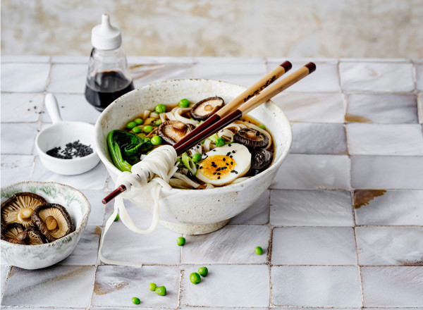 Eine Schüssel mit leckerer Nudelsuppe, garniert mit einem weich gekochten Ei, frischen Erbsen, Gemüse und Pilzen. Zwei Essstäbchen liegen auf der Schüssel. Nebenan befindet sich eine kleine Schale mit getrockneten Pilzen.